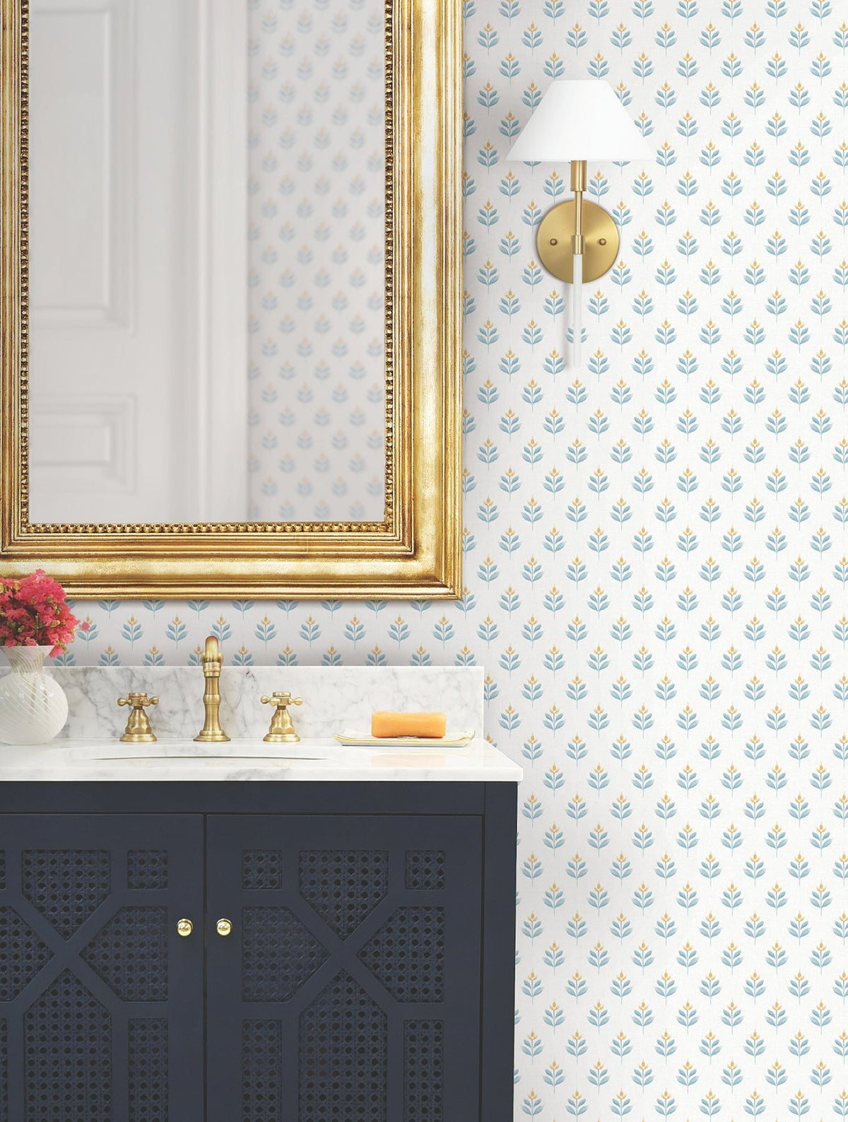 Bathroom with a gold-framed mirror, Serene Villa Floral Stamp Wallpaper by York Wallcoverings, wall sconce, dark vanity with marble top, brass faucet, red flowers in a vase, and a yellow soap on a tray.