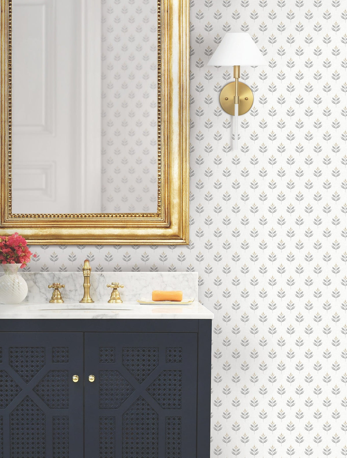 A bathroom vanity with a gold faucet, marble countertop, gold-framed mirror, wall sconce, soap bar, and a vase of flowers stands against a wall covered in Serene Villa Floral Stamp Wallpaper by York Wallcoverings—a white backdrop adorned with a delicate gray leaf pattern.