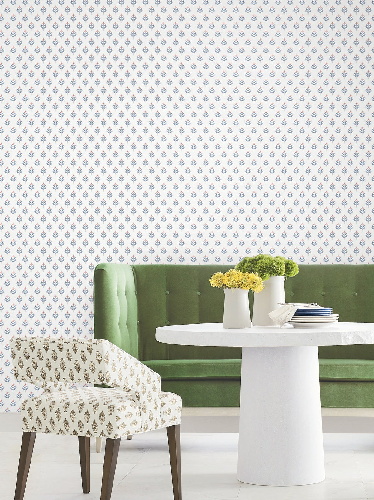 Sure! Here’s a rewritten version of the sentence using your product data: A green tufted sofa, a round white table adorned with flowers and books, and a patterned chair are arranged against a white wall covered in Serene Villa Floral Stamp Wallpaper by York Wallcoverings.