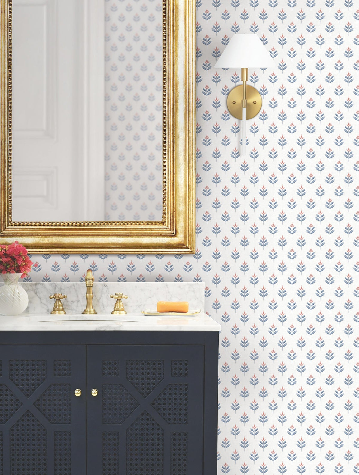 Bathroom with a gold-framed mirror, marble counter, black vanity, brass faucet, and a small vase of red flowers. The walls are adorned with Serene Villa Floral Stamp Wallpaper by York Wallcoverings, accompanied by a wall sconce. A yellow soap sits on the counter.