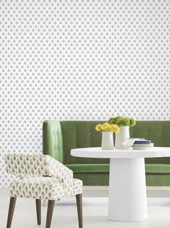 A green velvet sofa and a white round table adorned with white vases and yellow flowers are arranged in front of a wall featuring the Serene Villa Floral Stamp Wallpaper by York Wallcoverings, which displays a delicate blue and pink floral pattern. Next to the table sits a patterned chair with wooden legs.