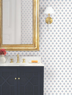Bathroom with a gold-framed mirror, brass faucet, marble countertop, dark blue vanity, York Wallcoverings’ Serene Villa Floral Stamp Wallpaper, wall sconce, and a small vase of red flowers. Soap rests on the counter.