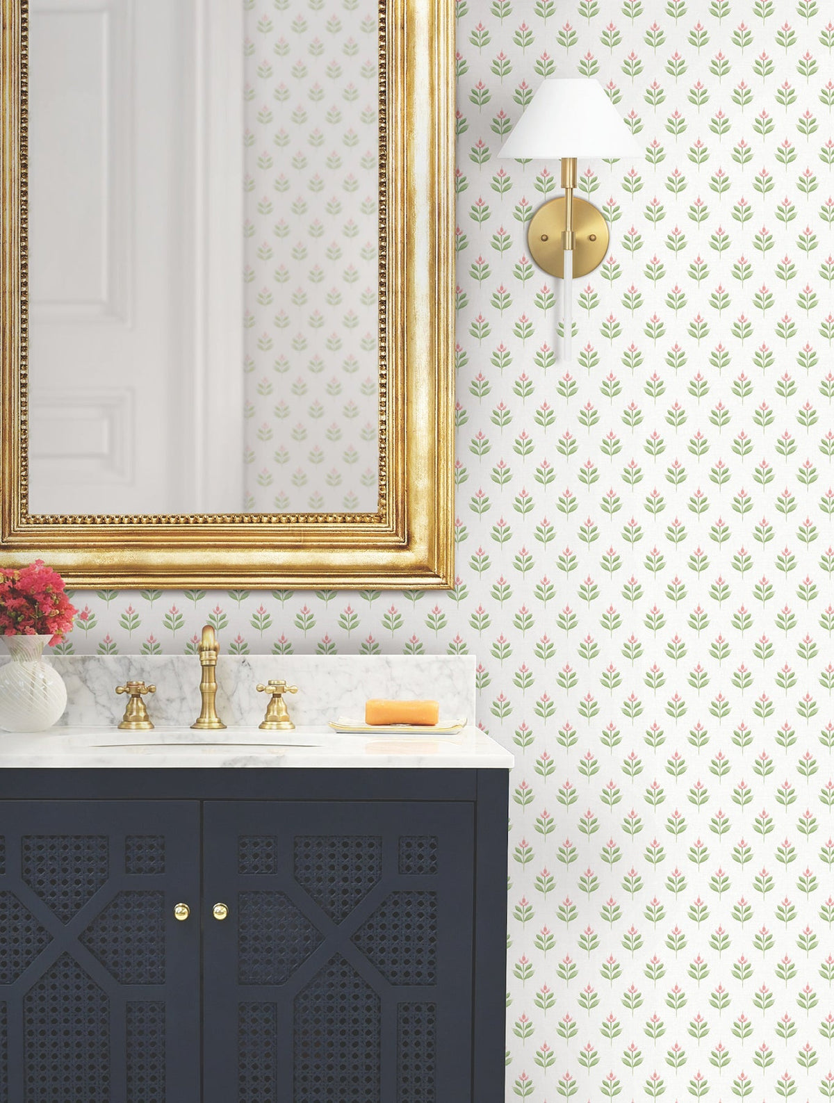 A bathroom scene featuring a gold-framed mirror, a wall sconce, a navy blue vanity with a marble countertop, brass fixtures, a soap bar, and a small vase with pink flowers is elevated by the Serene Villa Floral Stamp Wallpaper from York Wallcoverings.