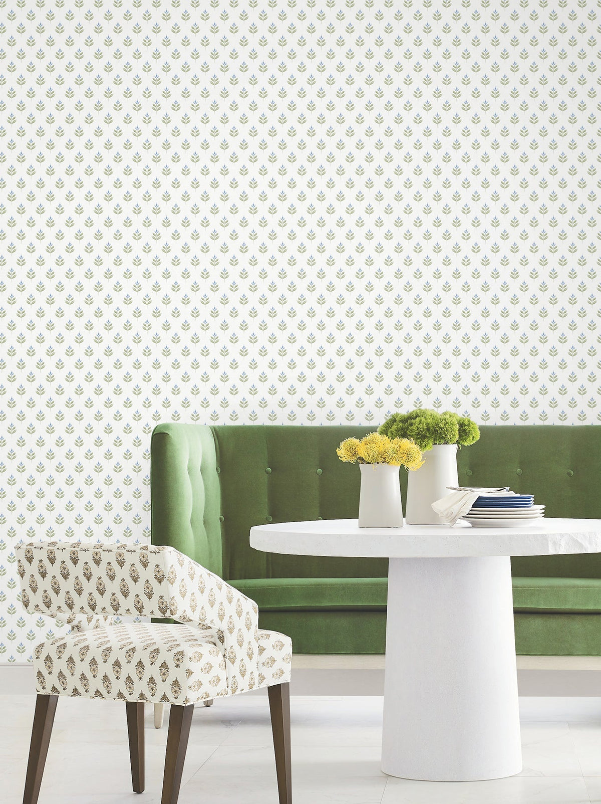 A green velvet banquette, white round table topped with yellow flowers in vases, and a patterned accent chair are featured in a modern, bright room with Serene Villa Floral Stamp Wallpaper by York Wallcoverings, showcasing a small green and yellow floral motif.