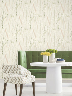 A white round table with yellow flowers, a green tufted bench, and a patterned chair sit in front of Meadow Splendor Botanical Wallpaper, featuring delicate grass and plant illustrations in soft green and beige tones.