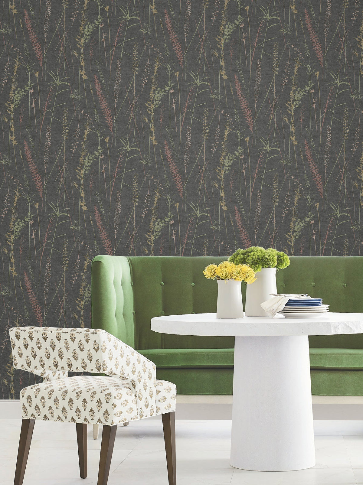 A green velvet tufted bench, a white round table adorned with yellow and green flowers, and a patterned chair sit in front of the Meadow Splendor Botanical Wallpaper.