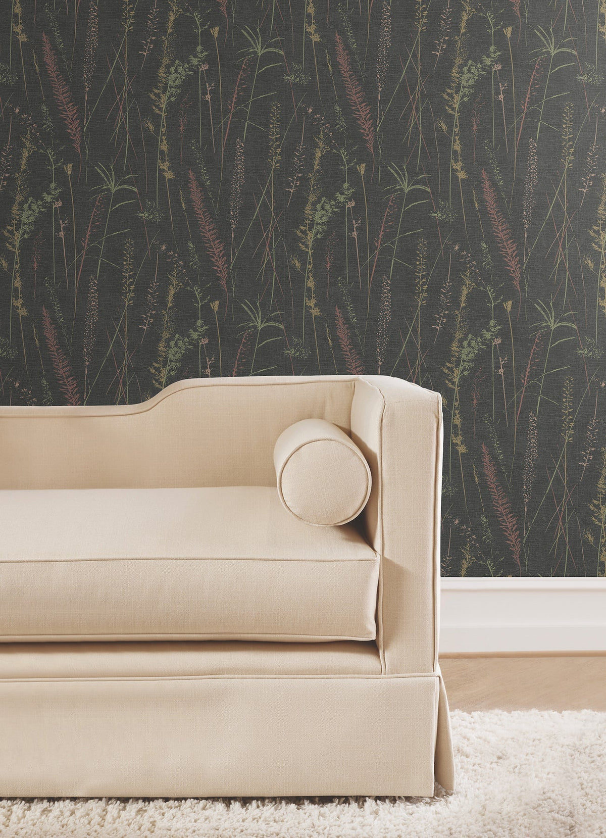 A beige couch with a round pillow sits on a tan rug against Meadow Splendor Botanical Wallpaper and a white baseboard.