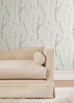A beige upholstered sofa with a round bolster pillow sits on a wooden floor in front of Meadow Splendor Botanical Wallpaper featuring a light botanical pattern with blue and green grasses.