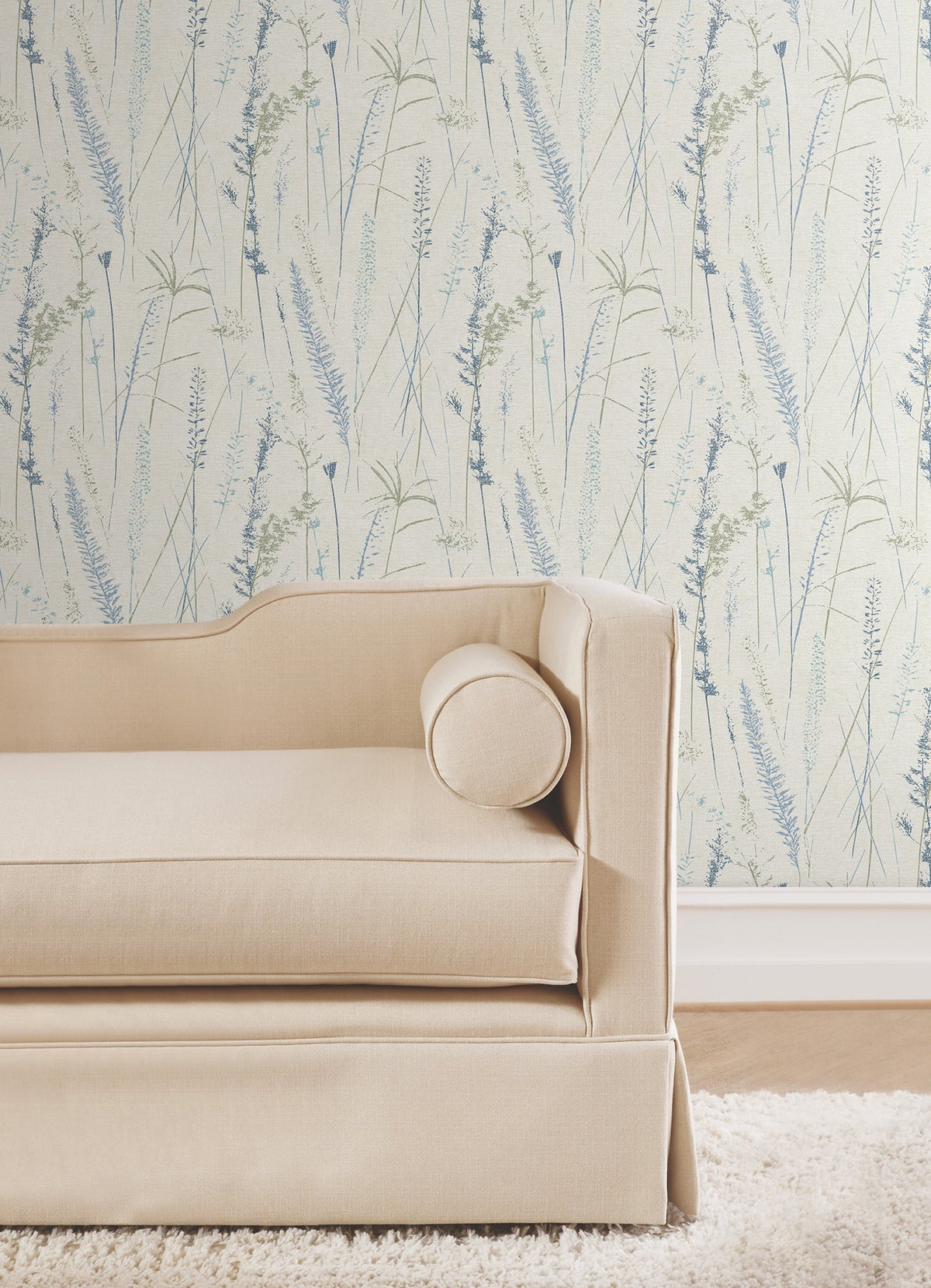 Beige upholstered sofa with a round bolster pillow against a light wall featuring the Meadow Splendor Botanical Wallpaper in delicate blue and green tones.
