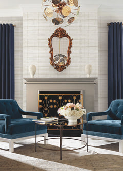Two blue velvet chairs face a glass coffee table with flowers, set before a fireplace with a gold floral screen. A decorative mirror and vases are on the mantel, framed by navy curtains and walls adorned in Vanishing Point Striated Wallpaper by York Wallcoverings.