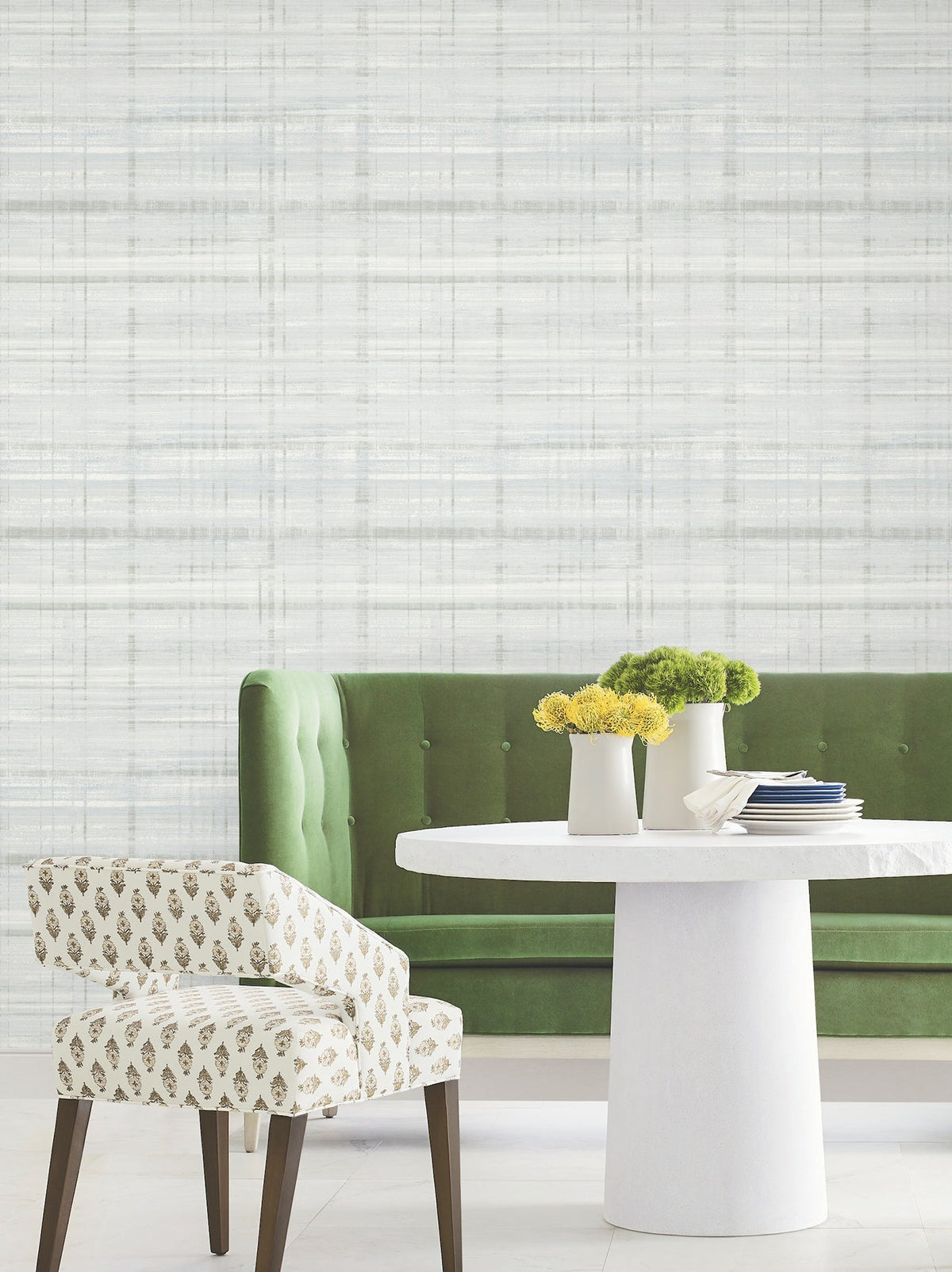 A green velvet sofa, a white round table with two white vases of yellow and green flowers, and a patterned chair set against York Wallcoverings’ Vanishing Point Striated Wallpaper.