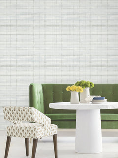 A green velvet sofa, a white round table with two white vases of yellow and green flowers, and a patterned chair set against York Wallcoverings’ Vanishing Point Striated Wallpaper.