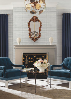 Elegant living room featuring Vanishing Point Striated Wallpaper by York Wallcoverings, two blue armchairs, a glass coffee table with a white flower arrangement, a decorated fireplace, dark curtains, two white vases, and a gold mirror above the mantel.