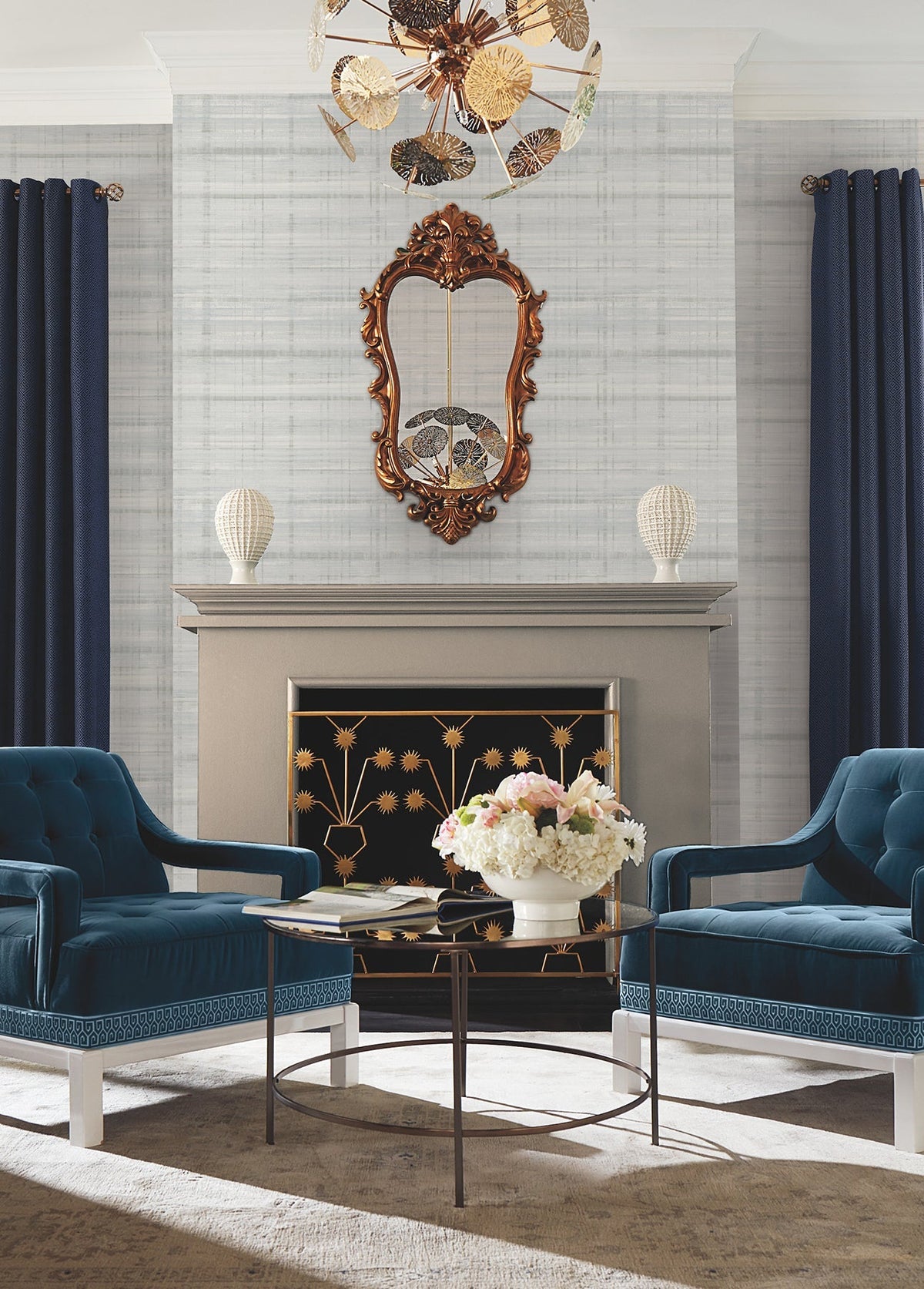 Certainly! Here’s a revised version of the sentence that incorporates your product data: Elegant living room featuring York Wallcoverings’ Vanishing Point Striated Wallpaper, two blue velvet chairs, a glass coffee table with white flowers, a fireplace with gold floral screen, ornate mirror above, white vases on the mantel, navy curtains, and a decorative ceiling light fixture.