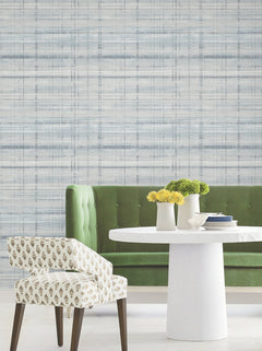 A green tufted bench, a round white table topped with yellow flowers in vases, and a patterned chair are arranged in front of York Wallcoverings’ Vanishing Point Striated Wallpaper. The setting is modern and bright with minimal decor.