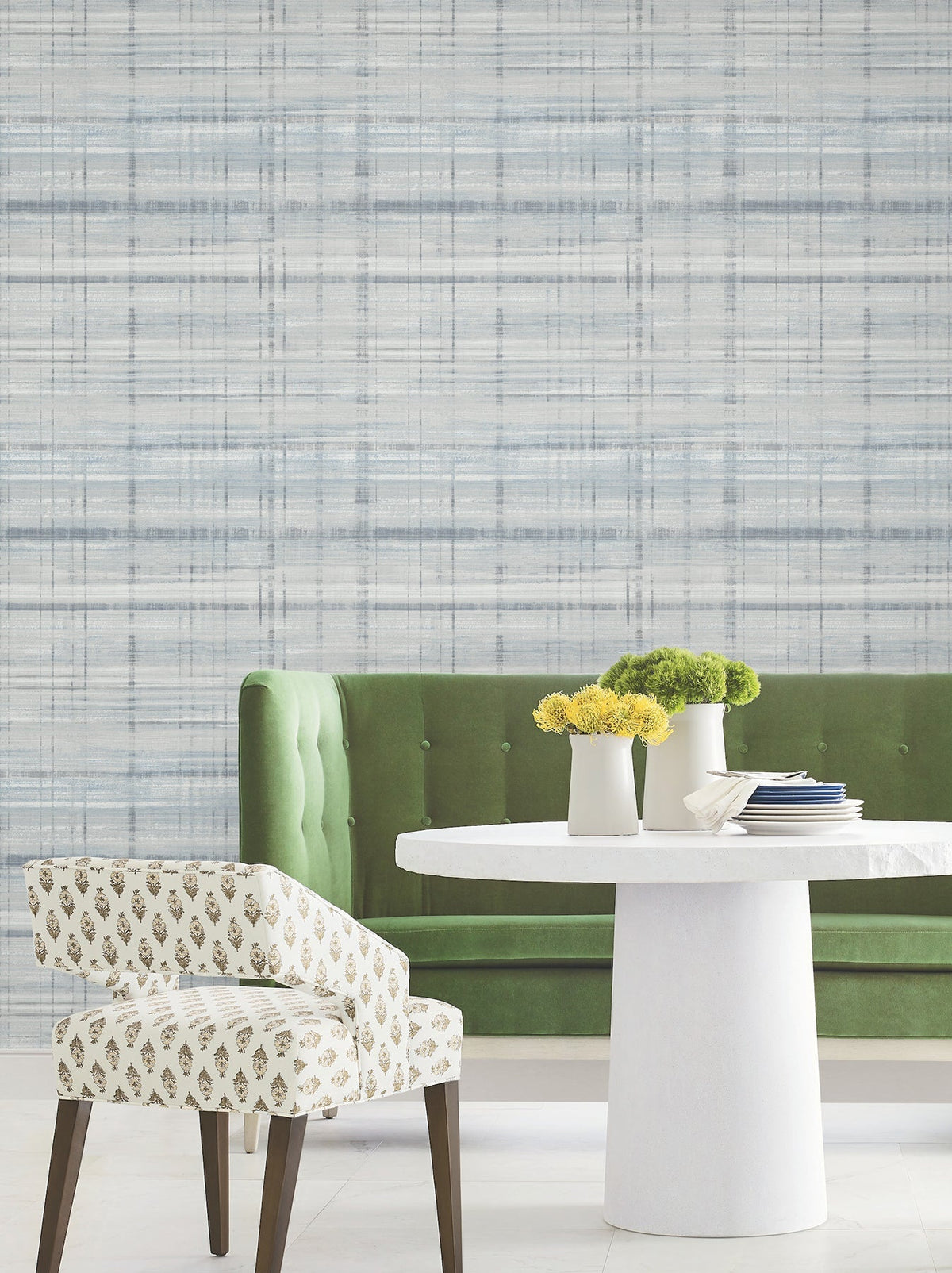 A green upholstered bench and a patterned chair sit by a round white table with small yellow flower vases, set against a blue and gray wall featuring York Wallcoverings’ Vanishing Point Striated Wallpaper.