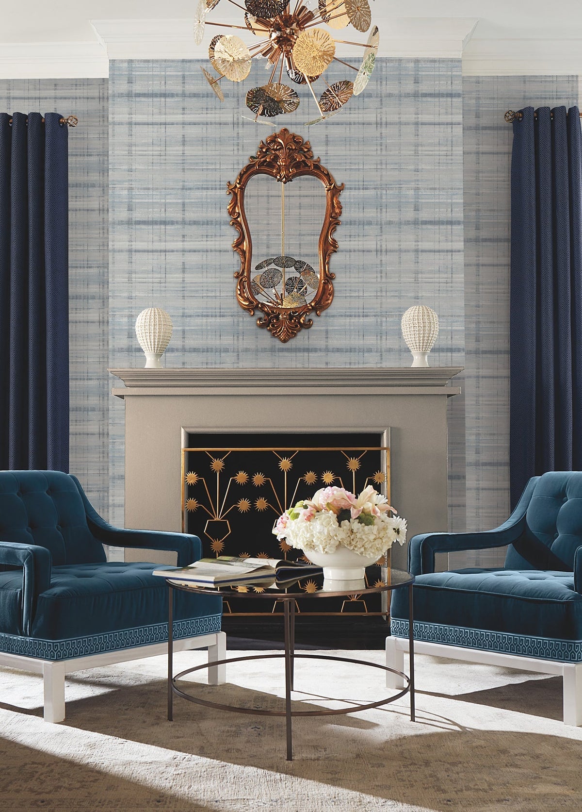 Elegant living room with blue armchairs, a modern coffee table topped with a white floral centerpiece, gold-accented fireplace, ornate mirror, blue drapes, and contemporary chandelier set against York Wallcoverings’ Vanishing Point Striated Wallpaper.