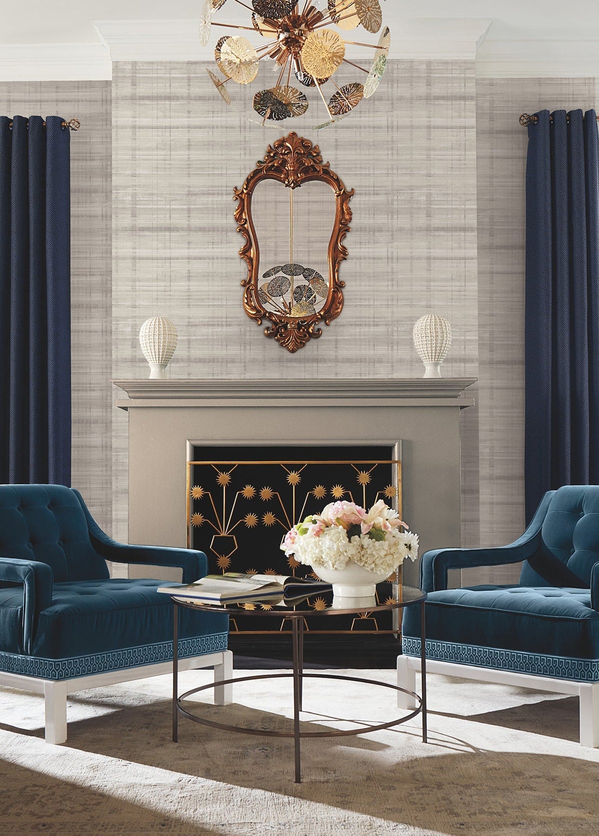 Two blue velvet chairs face a small table with a white flower arrangement, set in front of a fireplace. A decorative mirror hangs above the mantel against Vanishing Point Striated Wallpaper by York Wallcoverings, flanked by navy curtains and two textured white vases.