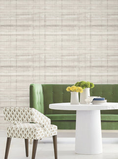 A green tufted sofa, a white round table with two white vases of yellow flowers, and a patterned upholstered chair sit against a wall covered in Vanishing Point Striated Wallpaper by York Wallcoverings.
