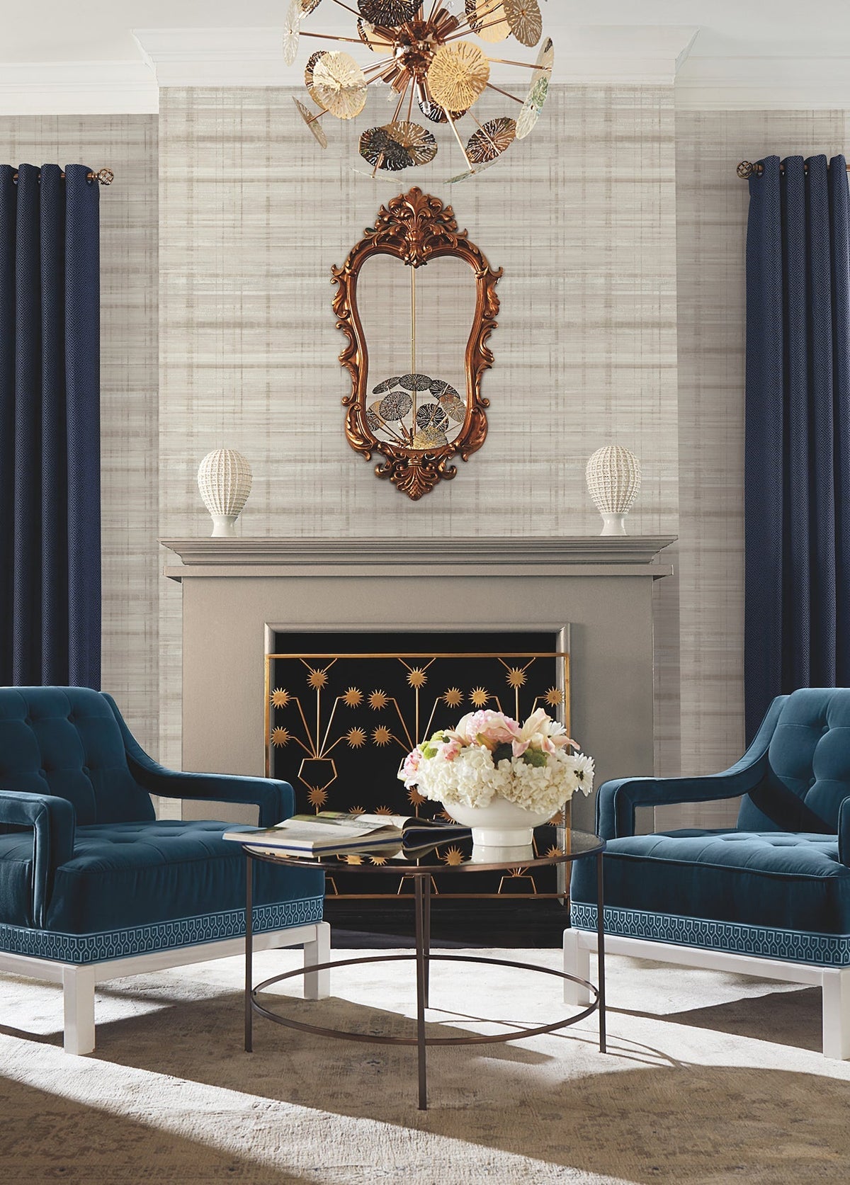 Two blue velvet chairs face a glass coffee table with flowers, set before a gray fireplace. Vanishing Point Striated Wallpaper by York Wallcoverings creates a refined backdrop, while a gold-framed mirror, ornate chandelier, and navy curtains complete the elegant living room decor.