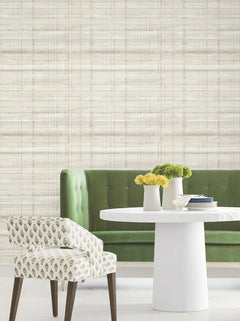 A modern dining area featuring a green tufted bench, a round white table, a patterned white chair, and two vases with yellow and green flowers is set against a beige Vanishing Point Striated Wallpaper by York Wallcoverings.