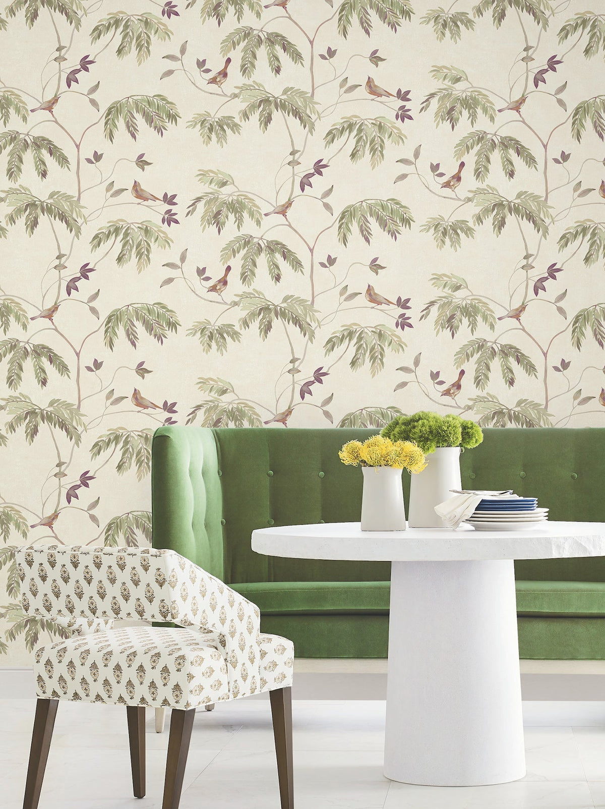 A green tufted sofa, a round white table with yellow and green flowers, and a patterned chair sit in front of York Wallcoverings’ Cillian Botanical Bird Trail Wallpaper.
