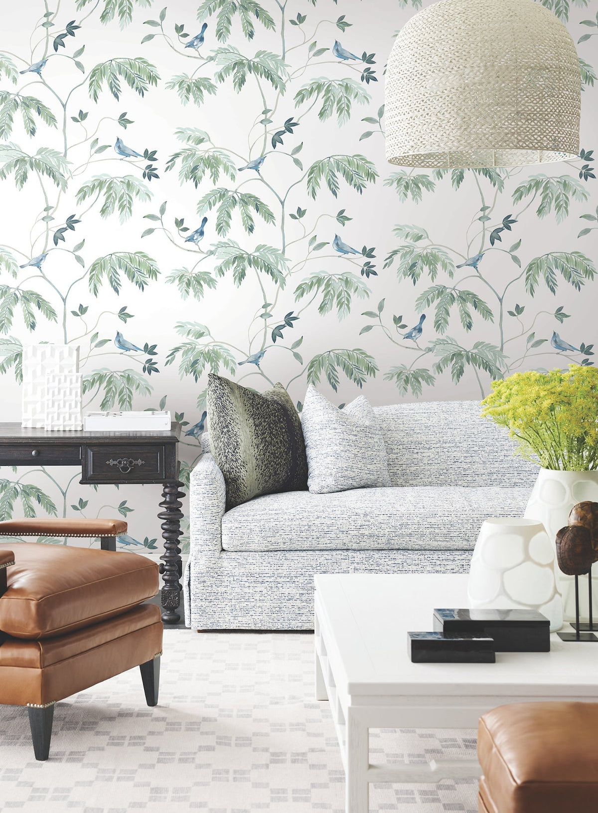 Living room featuring the Cillian Botanical Bird Trail Wallpaper by York Wallcoverings, a patterned sofa, leather chair, black side table, white coffee table, modern decor accents, a large pendant light, and a vase of yellow flowers on the table.