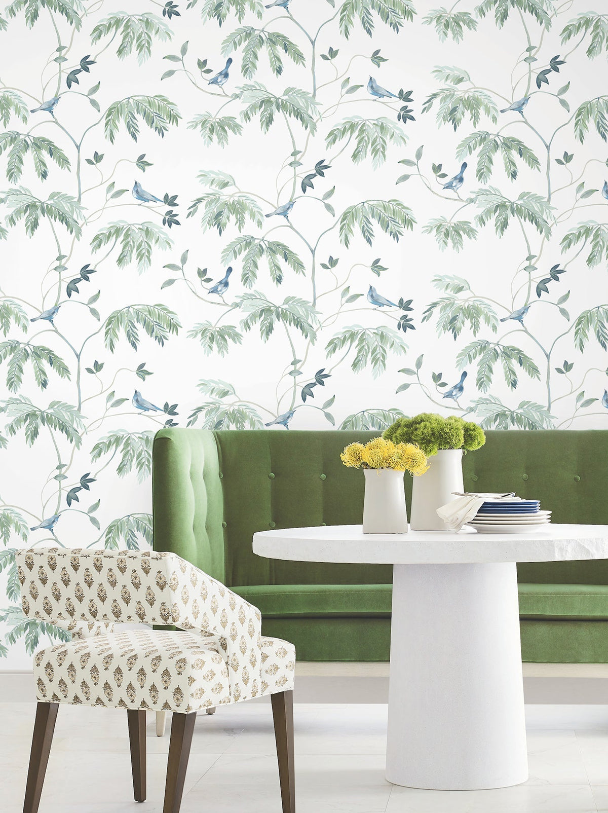 A white table with yellow flowers, a green velvet bench, and a patterned chair sit in front of a wall adorned with Cillian Botanical Bird Trail Wallpaper by York Wallcoverings.