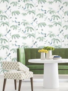 A white table with yellow flowers, a green velvet bench, and a patterned chair sit in front of a wall adorned with Cillian Botanical Bird Trail Wallpaper by York Wallcoverings.