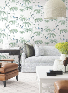 A bright living room with York Wallcoverings’ Cillian Botanical Bird Trail Wallpaper, a light sofa with pillows, a black side table, a white coffee table, a brown leather armchair, a vase of yellow flowers, and a woven pendant lamp.