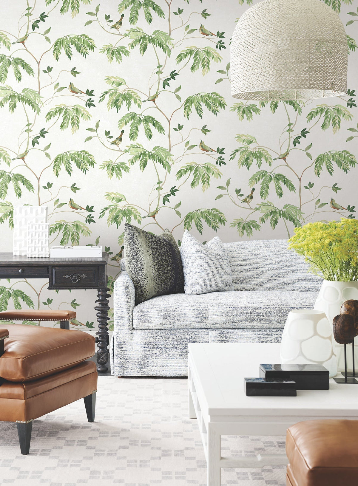 A stylish living room with York Wallcoverings’ Cillian Botanical Bird Trail Wallpaper, a light blue sofa, leather armchair, black side table, white coffee table, yellow flowers, and a large textured hanging lamp.