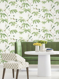 A green tufted sofa and a round white table with yellow flowers sit in front of York Wallcoverings’ Cillian Botanical Bird Trail Wallpaper. An upholstered chair with patterned fabric is positioned nearby, creating a bright and stylish scene.