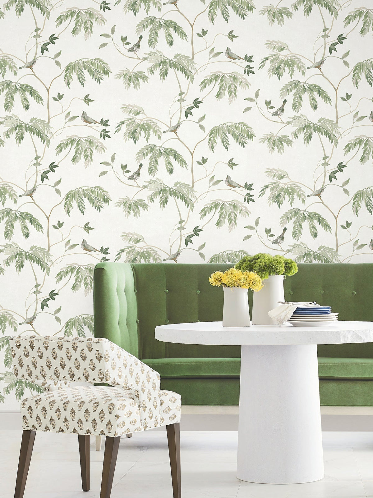A stylish room featuring a green tufted sofa, a white round table with vases of yellow flowers, a patterned chair, and leafy walls adorned in soft green and beige tones with the Cillian Botanical Bird Trail Wallpaper by York Wallcoverings.