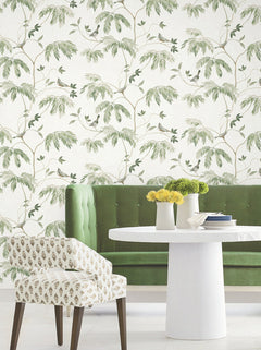 A green velvet sofa, a white round table adorned with yellow flowers in vases, and a patterned chair sit in front of the Cillian Botanical Bird Trail Wallpaper by York Wallcoverings, creating a light and airy atmosphere.
