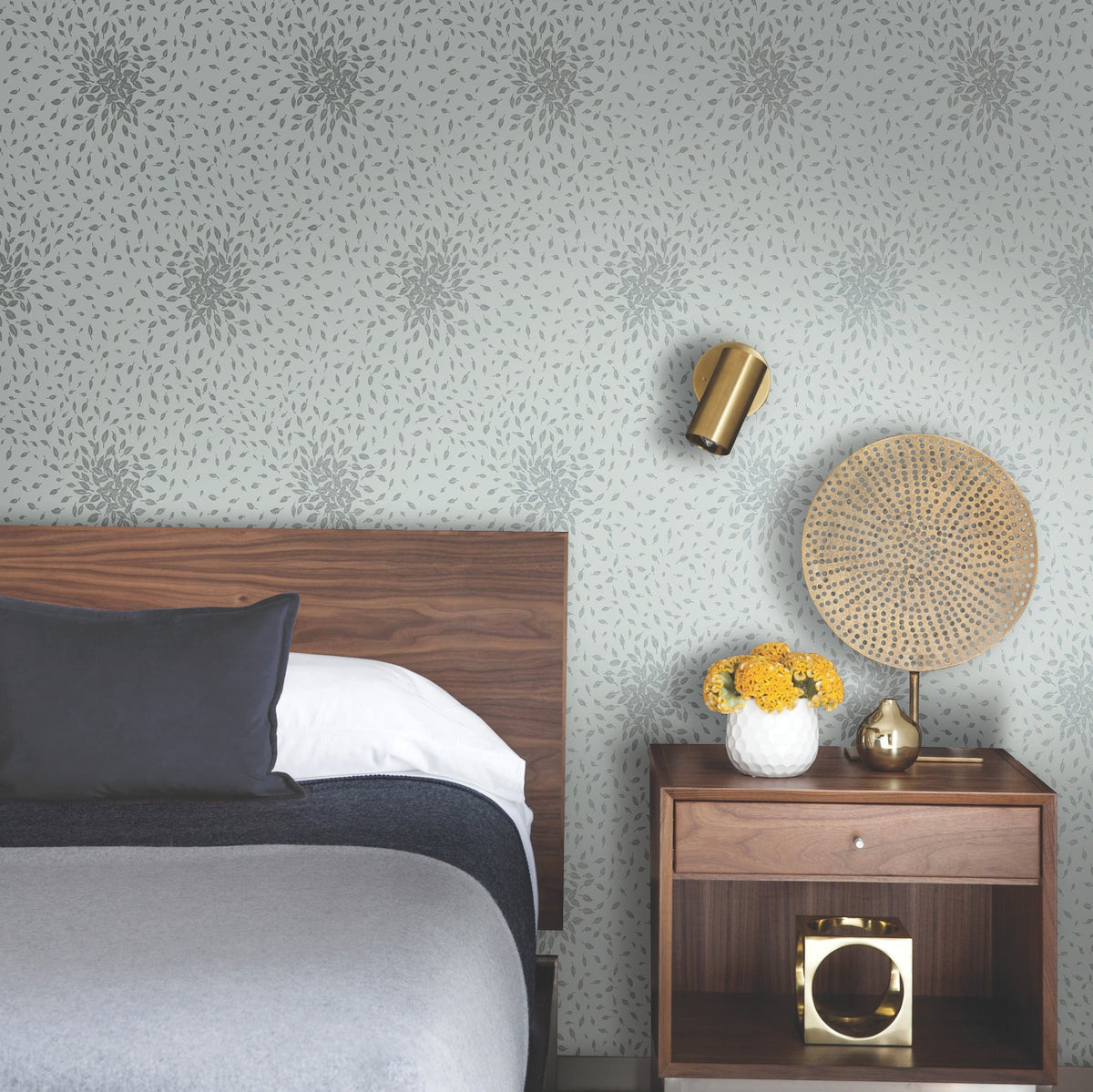 Modern bedroom with a wooden headboard, gray bedding, Petite Leaves Wallpaper by York Wallcoverings, a brass wall sconce, and a nightstand holding a vase of yellow flowers, a golden decorative object, and a woven fan.