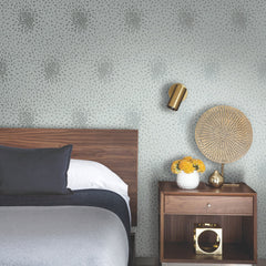 Modern bedroom with a wooden headboard, gray bedding, Petite Leaves Wallpaper by York Wallcoverings, a brass wall sconce, and a nightstand holding a vase of yellow flowers, a golden decorative object, and a woven fan.