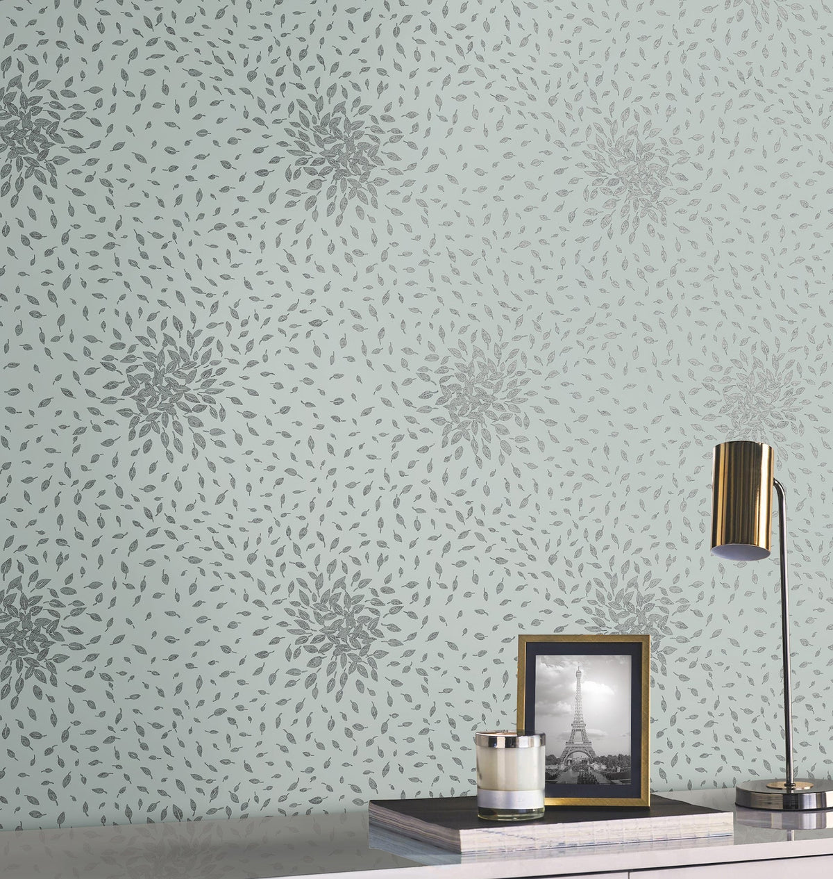 A desk with a gold lamp, a framed Eiffel Tower photo, a candle, and a book sits in front of a light blue wall adorned with York Wallcoverings’ Petite Leaves Wallpaper.