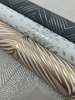 Three rolls of York Wallcoverings Petite Leaves Wallpaper featuring metallic designs—dark grey with silver lines, light grey with silver leaves, and white with gold and silver abstract lines—rest on patterned paper adorned with silver dots and lines.