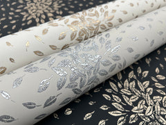 Three rolls of York Wallcoverings’ Petite Leaves Wallpaper, featuring metallic gold and silver leaf patterns on black and white backgrounds with coordinating leaf designs.