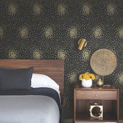 A modern bedroom featuring York Wallcoverings’ Petite Leaves Wallpaper in a dark floral motif, complemented by a wooden headboard and nightstand. Décor highlights include a gold wall sconce, a vase with yellow flowers, round woven wall art, and elegant gold accents.