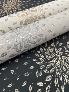 Three rolls of York Wallcoverings’ Petite Leaves Wallpaper, each featuring metallic leaf patterns in silver, gold, and bronze tones, are displayed on a surface adorned with a matching gold leaf design set against a dark background.