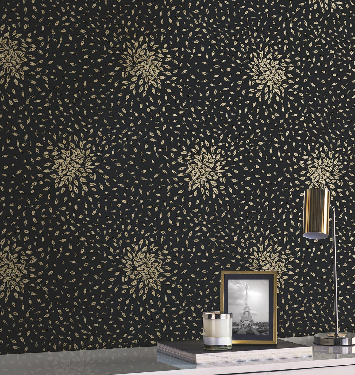 A desk with a gold lamp, a framed Eiffel Tower photo, and a cup sits against a dark wall adorned with Petite Leaves Wallpaper by York Wallcoverings.