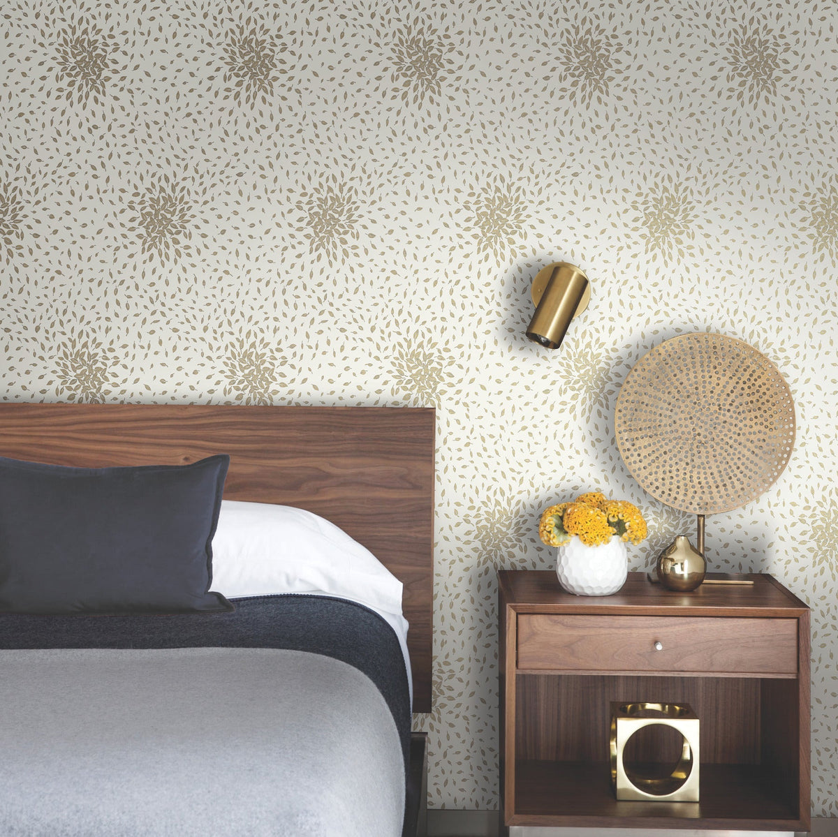 Modern bedroom with a wooden headboard, grey bedding, a nightstand with a woven lamp, gold wall sconce, a vase of yellow flowers, and Petite Leaves Wallpaper by York Wallcoverings.