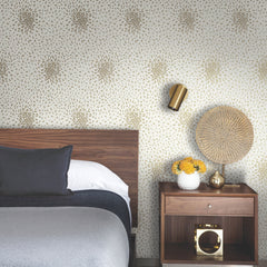 Modern bedroom with a wooden headboard, grey bedding, a nightstand with a woven lamp, gold wall sconce, a vase of yellow flowers, and Petite Leaves Wallpaper by York Wallcoverings.