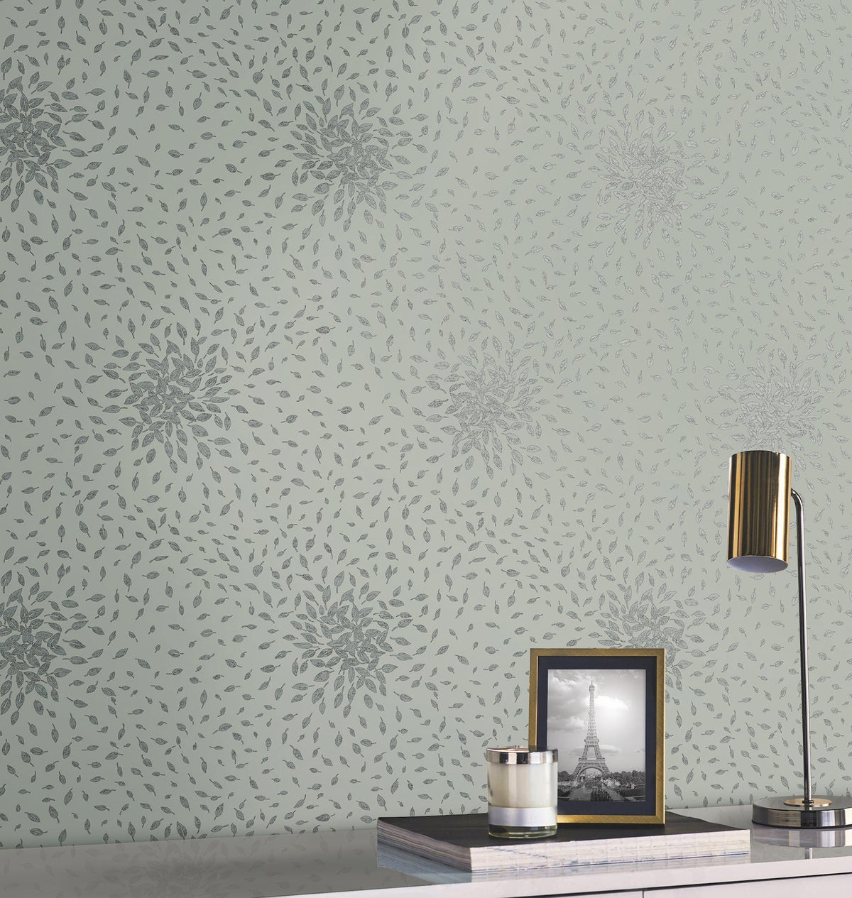 A wall decorated with York Wallcoverings’ Petite Leaves Wallpaper in a gray floral burst pattern on pale green sets the backdrop for a white desk, which displays a gold lamp, a glass candle, and a framed black and white photo of the Eiffel Tower.