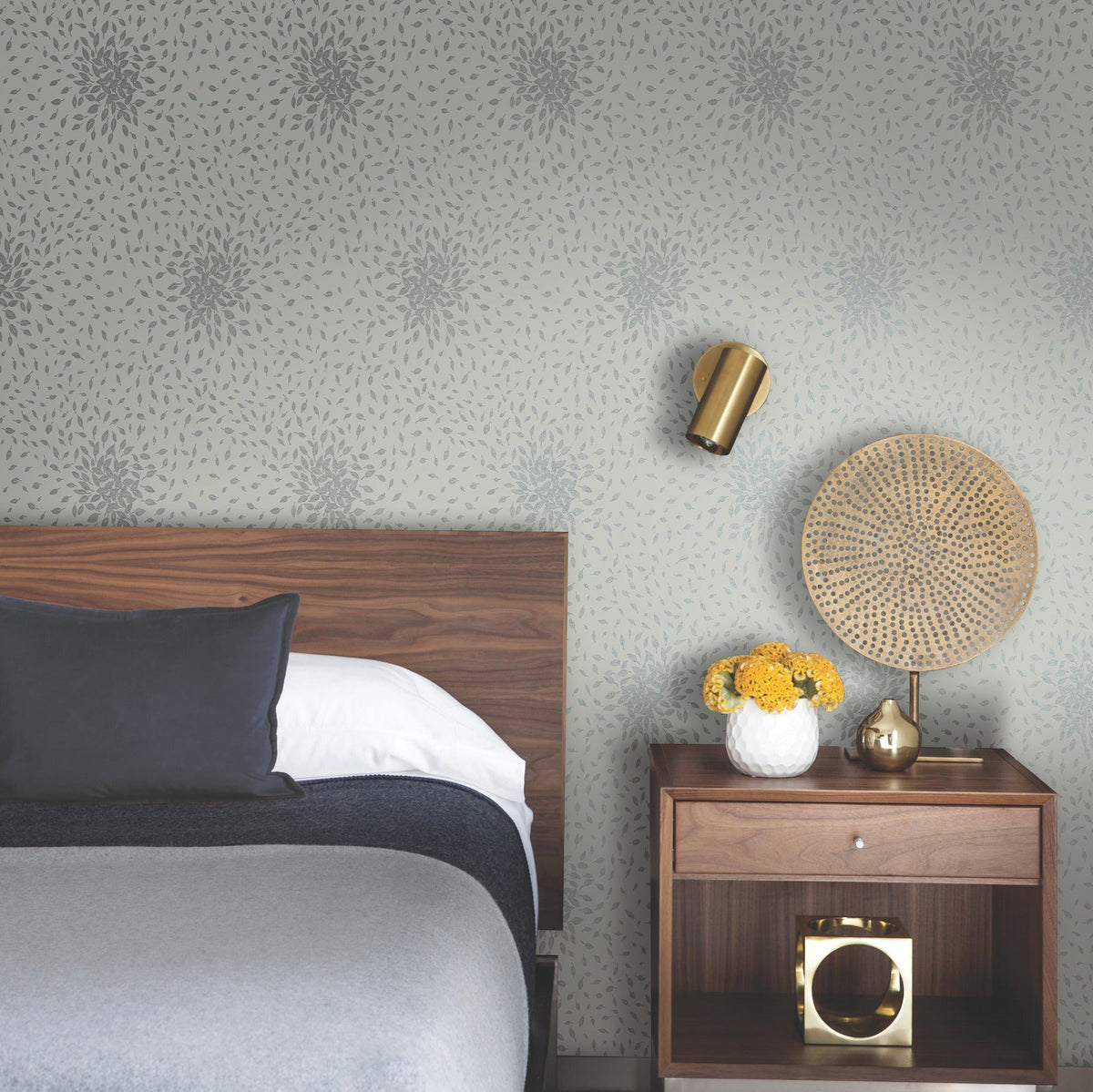 A modern bedroom featuring the Petite Leaves Wallpaper by York Wallcoverings as a backdrop, with a wooden headboard, gray bedding, a nightstand topped with a vase of yellow flowers, a gold table lamp, and a round wicker decor piece.