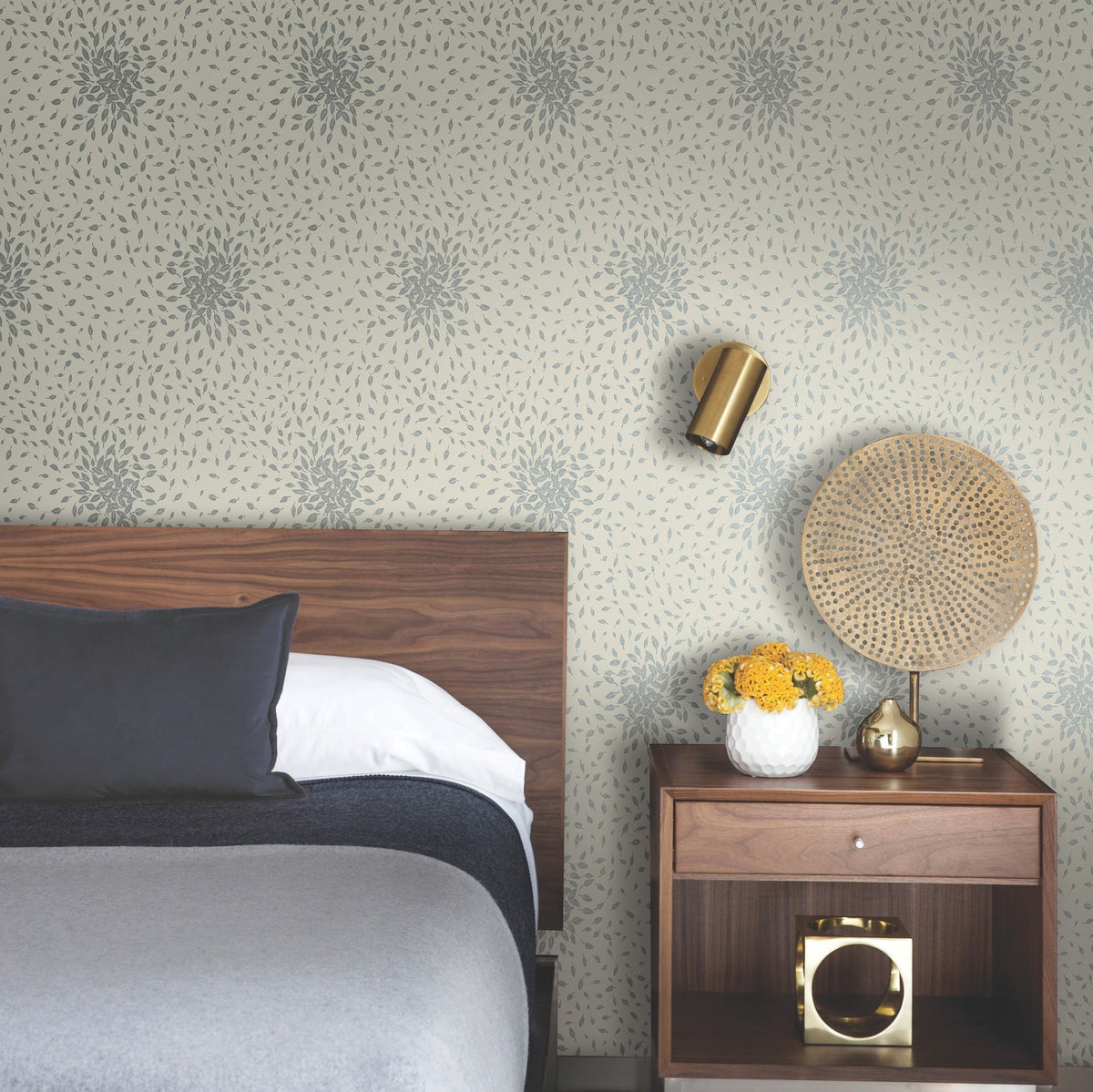 A modern bedroom with a wooden headboard, gray bedding, Petite Leaves Wallpaper by York Wallcoverings, a nightstand holding a vase of yellow flowers, a golden lamp, and a decorative round wicker piece.