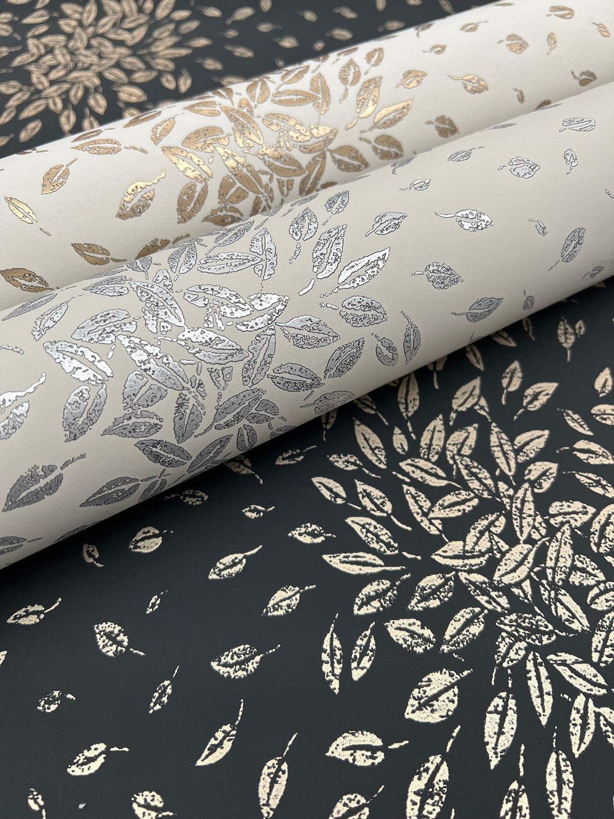 Three rolls of York Wallcoverings’ Petite Leaves Wallpaper, showcasing metallic leaf patterns in gold and silver, rest on a black surface adorned with similar leafy motifs. The design evokes a refined and nature-inspired aesthetic.