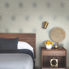 A modern bedroom featuring York Wallcoverings’ Petite Leaves Wallpaper, a wooden headboard, gray bedding, a nightstand with a wicker lamp, a gold vase with yellow flowers, and a gold wall sconce.