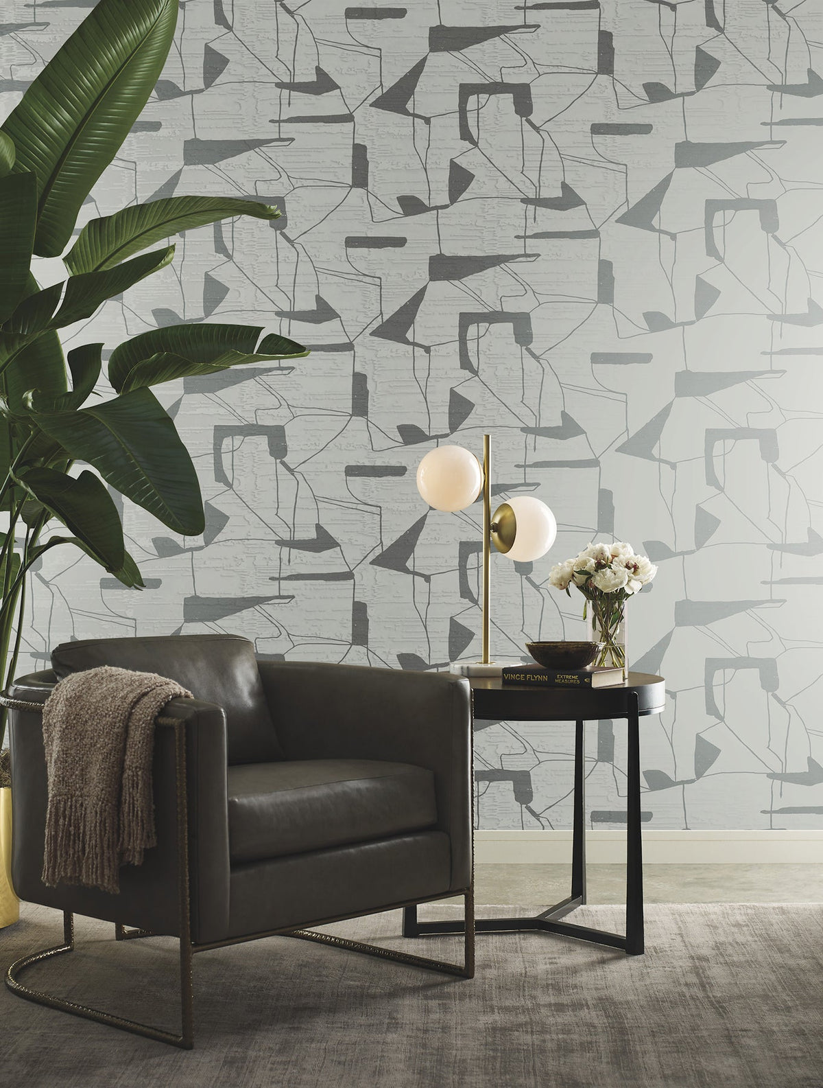 A modern living space featuring a gray armchair, a round black side table with a lamp and flowers, a large leafy plant, and York Wallcoverings’ Abstract Geo Wallpaper in the background.